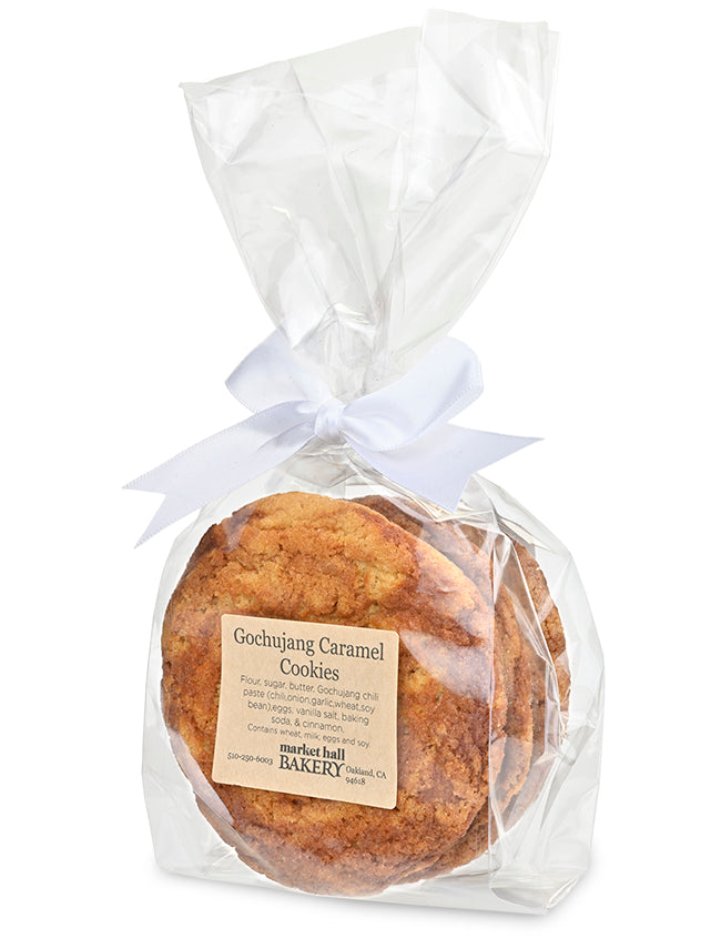 A packaged loaf of Gochujang Caramel Cookies from Market Hall Bakery, wrapped in clear cellophane with a white ribbon bow and a beige product label, on a white background.