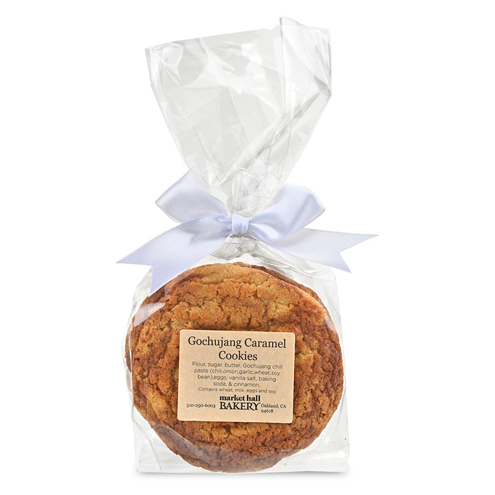 A packaged loaf of Gochujang Caramel Cookies from Market Hall Bakery, wrapped in clear cellophane with a white ribbon bow and a beige product label, on a white background.