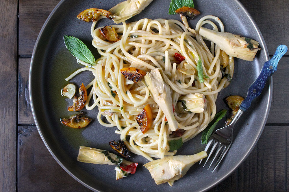 Plated dish of pasta wit artichokes, and charred lemon and mint on a wooden table.