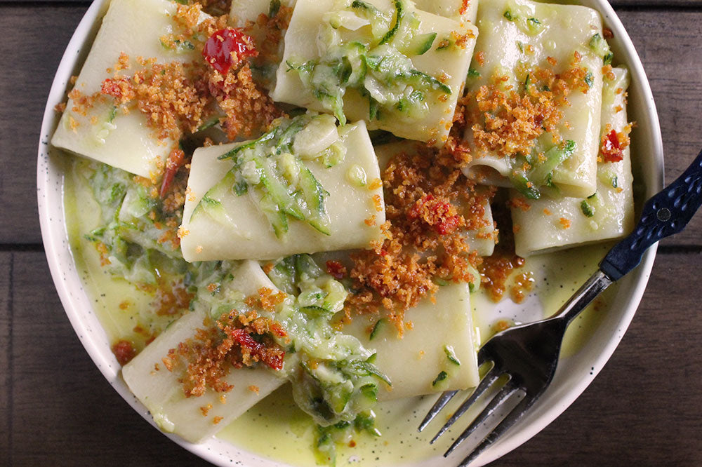 A close-up overhead shot of a bowl of pasta, likely rigatoni or similar hollow tubes, filled with a light green herb and cheese sauce, topped with breadcrumbs and red chili flakes, with a fork resting in the bowl on a dark wooden surface.