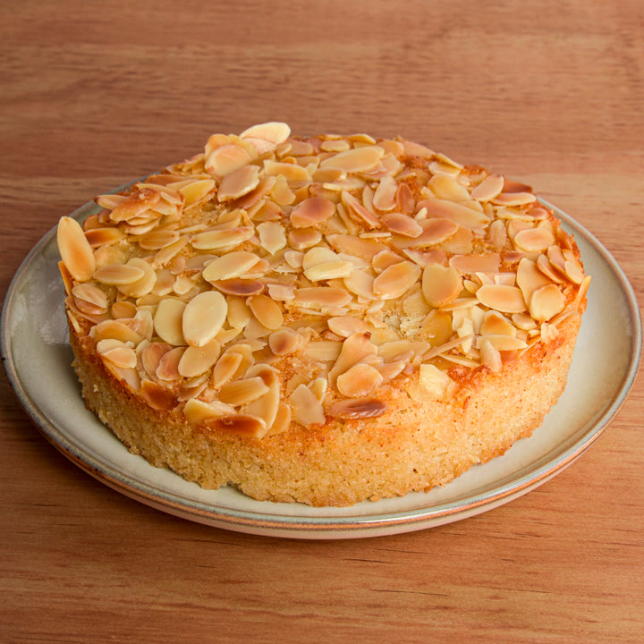 A round almond cake topped with sliced almonds on a ceramic plate, set on a wooden table.